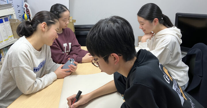 打合せ室にいる4人のメンバーの写真。奥の3人はスマホを手に話し合い、手前のメンバーが紙にメモしている。