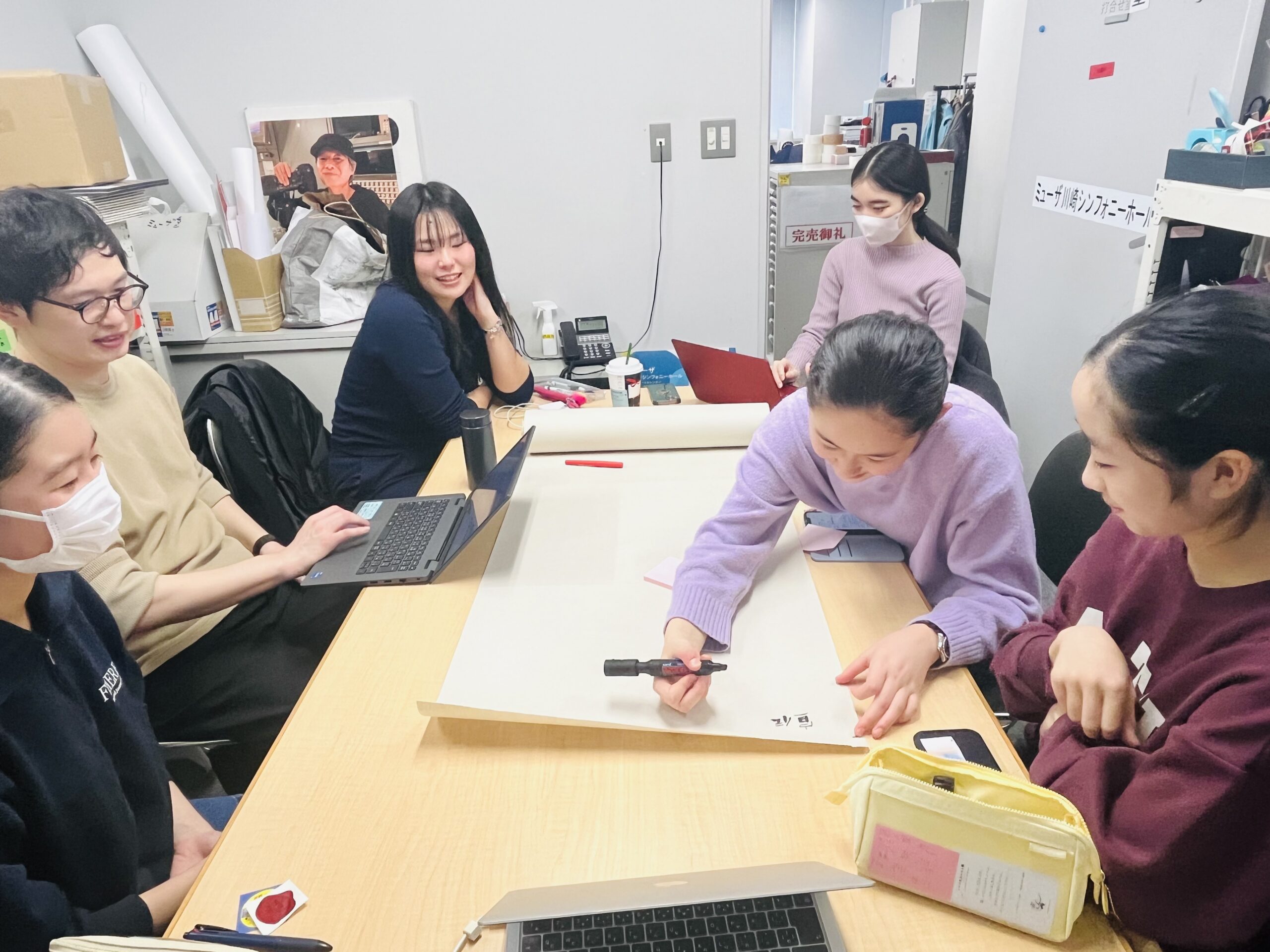 机に敷いた模造紙を中心に3人ずつ左右に座っている。右手に見える3人の中の真ん中の一人が黒いマジックペンを持ち、文字を書きはじめている。