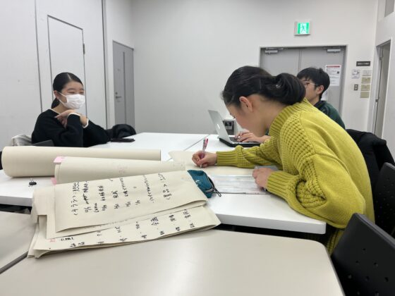 企画展示室でのミーティング風景。黄色のセーターを着たメンバーが資料に書き込みをしており、机の上にはメモ書きされた大きな用紙や書類が広げられている。