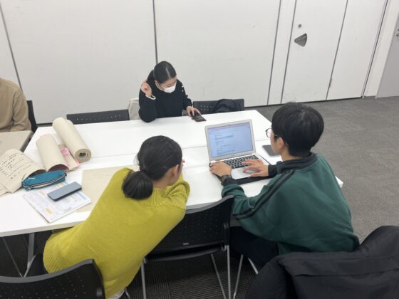 企画展示室で、3人のメンバーがノートパソコンや資料、巻紙を広げて作業に取り組んでいる様子。男性がパソコンを操作し、他のメンバーがその内容をのぞき込んでいる。