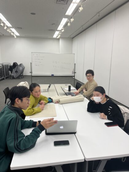 企画展示室でメンバーがテーブルを囲んでディスカッションをしている様子。手前のメンバーがノートパソコンの画面を指して発言しており、奥にはホワイトボードがある。