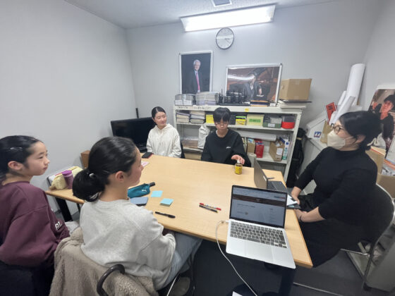 打合せ室の手前に座り、メンバーに話す山田さんと、山田さんの方を見て話を聞くメンバー4人の写真。