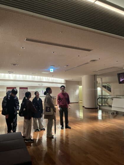 明かりが少しだけ着いているホールのホワイエを見渡している4人のメンバーと1人の大人スタッフの写真。