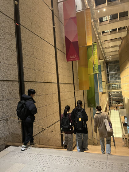 ホールのあるミューザビル4階から階段を降りる4人のメンバーの後ろ姿の写真。天井からはピンクや黄色、緑の大きなタペストリーが下げられている。