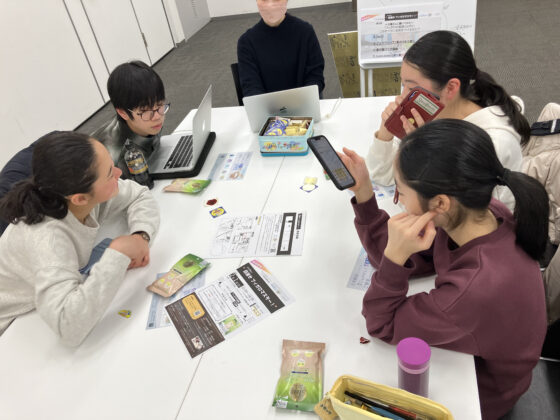 プリントや入浴剤が広げられた机を囲んで、パソコンやスマートフォンを手にしながら話し合っている様子の4人のメンバーの写真