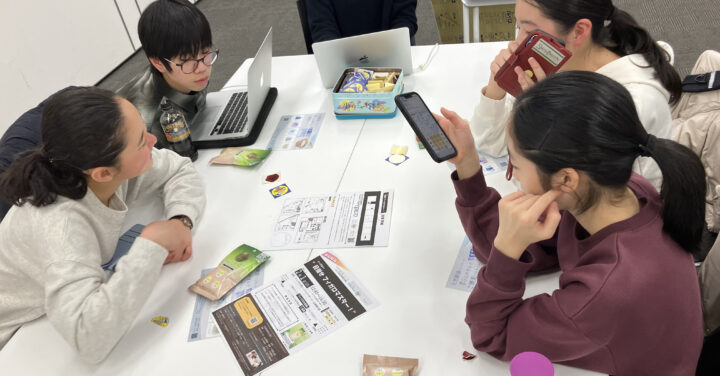 プリントや入浴剤が広げられた机を囲んで、パソコンやスマートフォンを手にしながら話し合っている様子の4人のメンバーの写真