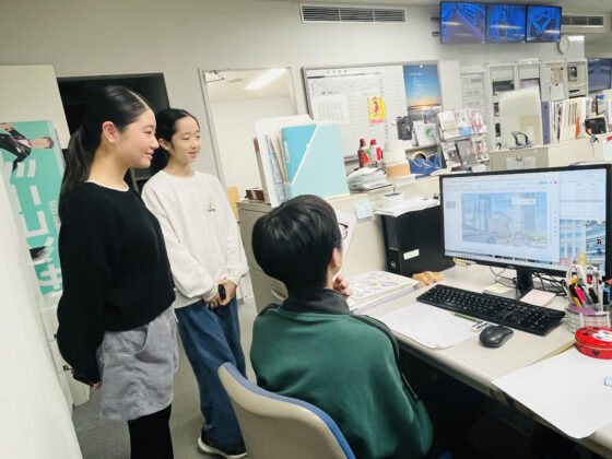 事務所でパソコンの画面を囲んで話し合う3人のメンバー。1人は椅子に座り、2人はその横に立って画面を注視している。