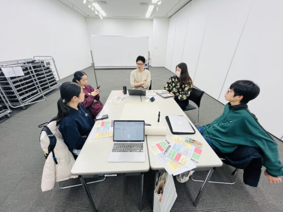 企画展示室で長机を囲み、3人のメンバーと2人の大人スタッフが話し合っている様子の写真。机上にはパソコンやカラフルなメモが広げられている。