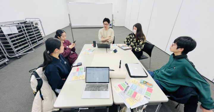 企画展示室で長机を囲み、3人のメンバーと2人の大人スタッフが話し合っている様子の写真。机上にはパソコンやカラフルなメモが広げられている。