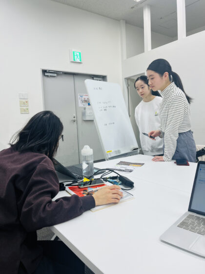 大人スタッフが座って机上のPCを経っているメンバー2人に見せている様子の写真