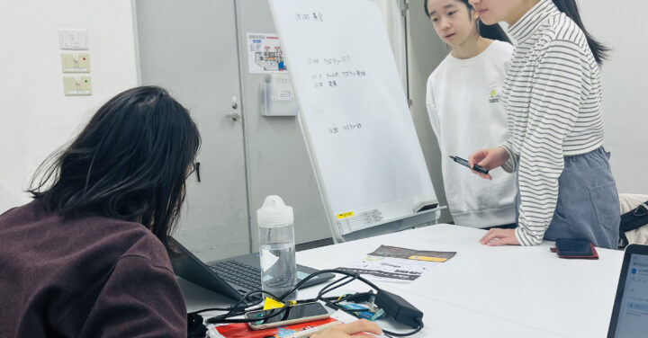 大人スタッフが座って机上のPCを経っているメンバー2人に見せている様子の写真