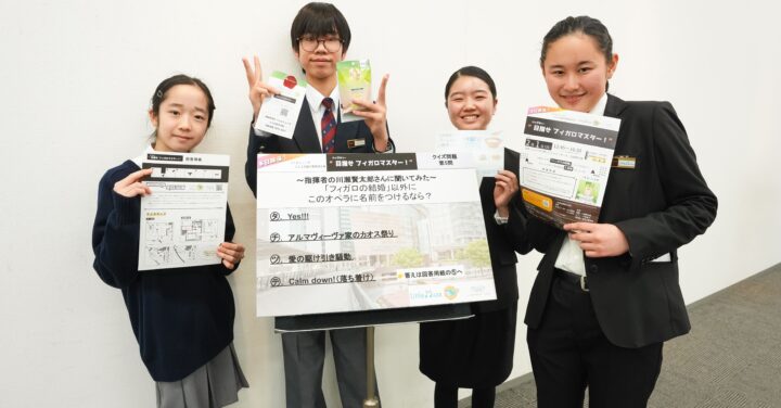 リトルミューザのメンバー4人が、各々チラシや解答用紙、景品の入浴剤を持ち、問題の掲示物を囲んでいる写真。笑顔や少し緊張した表情がうかがえる。