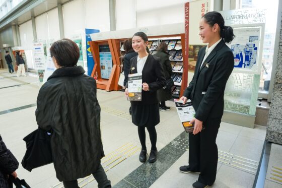 歓喜の広場でチラシを持って、お客様に緊張しながらも笑顔で声をかけるリトルミューザメンバー2人の写真