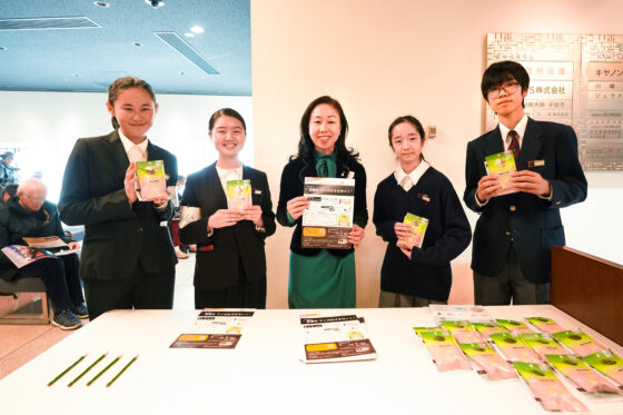 リトルミューザメンバー4人と株式会社しむらの社長様の集合写真。ホール入口付近の主催受付にて、メンバーは入浴剤を持ち、真ん中に立つ社長さまはチラシを持っている。