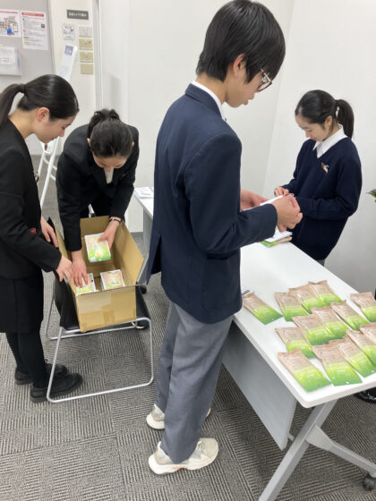 15個程の入浴剤パッケージが長机に並べられており、メンバーが広告の紙を貼る作業をしていたり、ダンボールに詰めたりしている様子の写真。入浴剤のパッケージは上が緑で下に向かってグラデーションで薄くなり、真ん中からしたはベージュの優しい色合い。