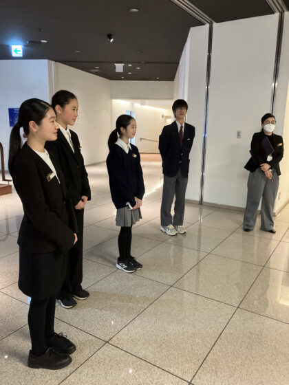 ホールのホワイエに立って並ぶリトルミューザのメンバー4人。通常活動時の普段着とは違い、スタッフとしてスーツや制服を着ている。横にはスタッフが立っている。