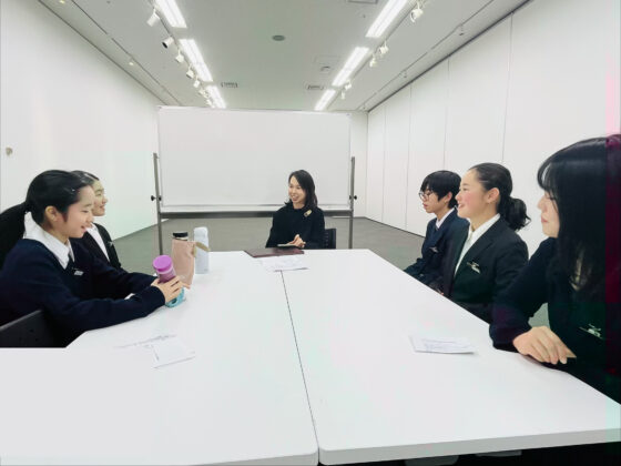 企画展示室で長机を囲んで話し合うメンバー4人と大学生アドバイザー、奥には大人スタッフがいる写真